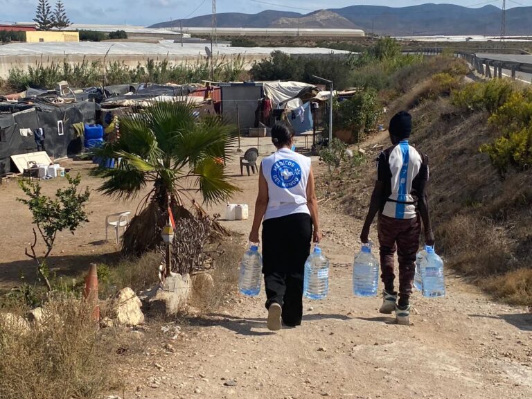 Trabajadora de Médicos del Mundo lleva agua potable en garrafas a un asentamiento informal en Almería junto a uno de los hombres que sobreviven en ese lugar.